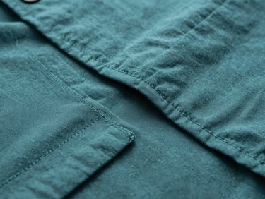 Close-up overhead view of housekeeping uniform fabric — durable cotton blend in muted teal, showing weave density and a reinforced seam under bright daylight-balanced studio light, no people