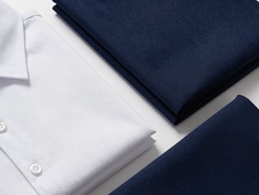Overhead flat-lay of neatly folded school uniform fabric — cotton-poly blend in white and navy, visible weave texture under bright even studio light, no people