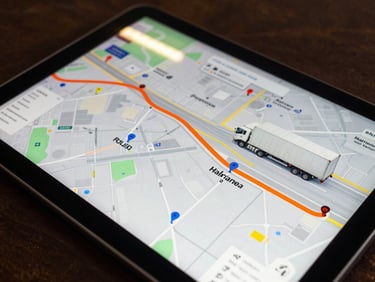 Close-up of a tablet screen showing a live GPS map with a truck route highlighted in orange, warehouse coordinates pinned, fluorescent warehouse lighting reflected on the screen surface