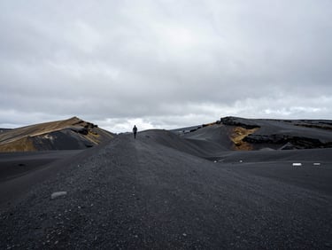 Ultra-wide environmental landscape of rugged Icelandic volcanic plains, a tiny lone figure walking along a black sand ridge under heavy overcast skies.