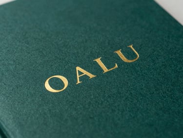 Close-up macro shot of a book cover with deep sea-green textured paper, minimalist gold foil typography, soft side-lighting with rich shadows.