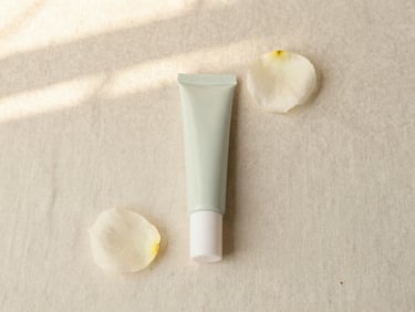 Flat lay of a clean beauty tinted balm tube and a single flower petal on a neutral linen backdrop, soft diffused light.