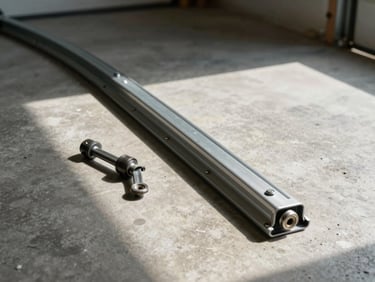 Overhead view of a bent garage door track section lying on a concrete garage floor next to replacement hardware, natural daylight casting hard shadows
