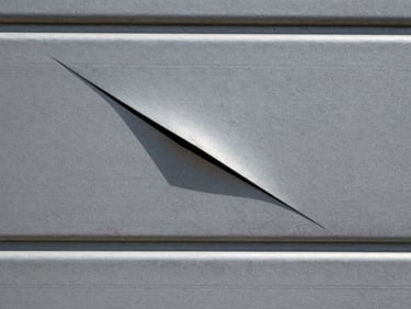 Close-up of a dented steel garage door panel, the crease running diagonally across the surface, natural outdoor light casting a shallow shadow across the damage