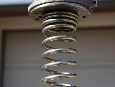 Extreme close-up of a coiled torsion spring mounted above a garage door, natural daylight, steel coils in sharp focus showing tension and wear