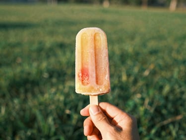 A hand holding a colorful, fruit-infused artisanal popsicle against a soft-focus green meadow backdrop, sun-drenched