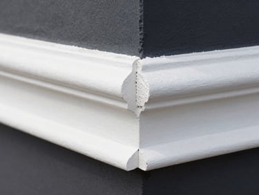 Macro shot of a perfect 45-degree miter joint on white architectural baseboard trim against a dark charcoal wall, high-contrast, sharp focus