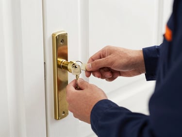 Close-up of a professional locksmith's hands inserting a shiny new key into a modern brass deadbolt on a white door, bright daylight.