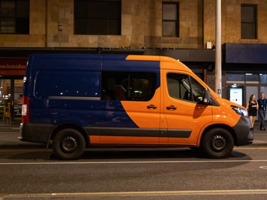 Wide environmental shot, warm street lighting at night, clean branded navy and orange van parked on a busy Melbourne street, crisp commercial photography, 35mm lens