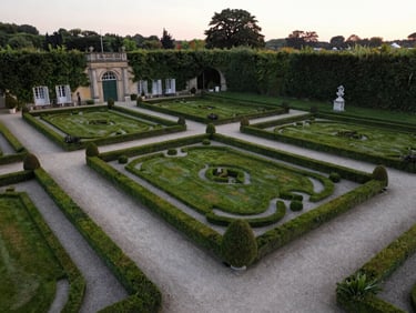 An aerial wide shot of a classic French estate garden with perfectly manicured hedges and gravel pathways, soft evening light.