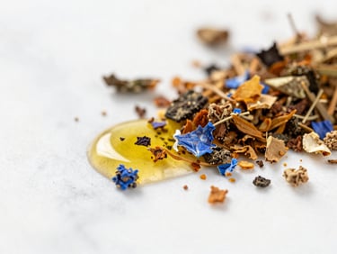Macro close-up of crushed raw botanicals and dripping golden oil on a white marble surface, bright natural daylight, soft focus.