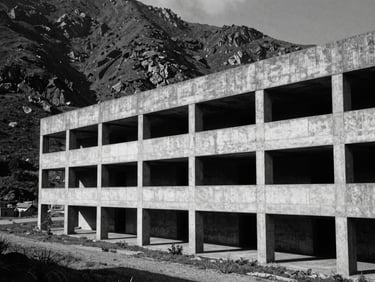 Wide establishing shot of a brutalist concrete structure cutting through a rugged mountain landscape, deep shadows, pure black and white, sharp geometric lines.