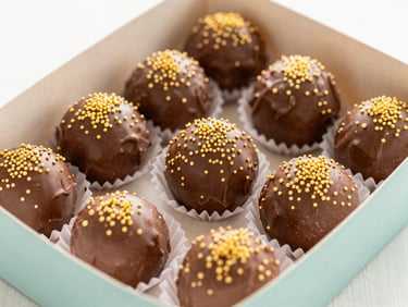Overhead shot of a box of glossy chocolate brigadeiros rolled in golden sprinkles, bright kitchen light