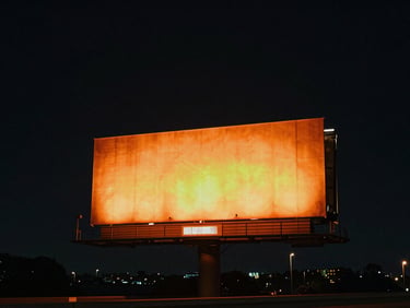 Wide environmental night shot of a colossal highway billboard glowing with vibrant orange light against a deep dark sky, cinematic atmosphere