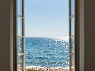 Interior of a light Normandy apartment, linen curtains half-open onto a grey sea horizon, bleached wood floor, morning overcast light, no people
