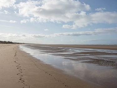 Wide environmental shot of the Normandy shoreline at low tide, overcast Nordic light, wet sand in foreground, horizon line low, empty beach stretching left, no people