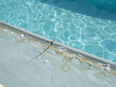 Close-up of a swimming pool interior surface showing active water loss damage — deep cracks in the render, calcium deposits along the waterline, surface blistering from years of Mediterranean UV and chemical exposure, overcast natural daylight, eye-level framing, no people