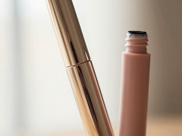Close-up of a sleek, modern mascara wand and tube, reflecting soft light, cream and blush tones, shallow depth of field