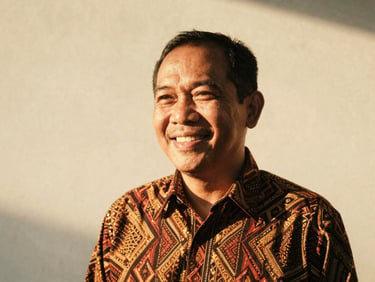 Candid portrait of a smiling Indonesian man in his 50s, warm natural golden-hour light, slightly faded analog tones, 35mm film aesthetic, wearing a batik shirt, looking friendly.
