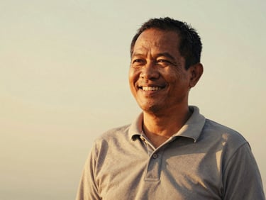 Candid portrait of a smiling Indonesian man in his 50s, warm natural golden-hour light, slightly faded analog tones, 35mm film aesthetic, wearing a casual polo shirt, looking relaxed and friendly.