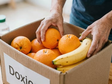 Candid close-up of hands placing fresh oranges and bananas into a cardboard donation box, bright morning light