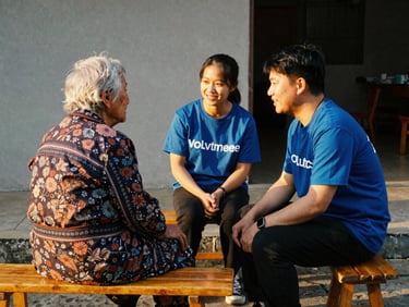 A volunteer sitting and chatting warmly with an elderly resident on a simple wooden bench outside, golden hour light