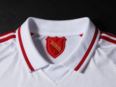 Detailed macro shot of a vintage white soccer jersey collar and embroidered red shield, dramatic side lighting, dark moody background, 50mm lens