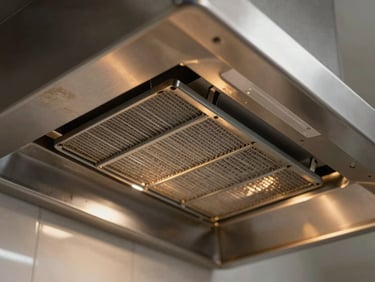Underside of a stainless steel kitchen hood extraction canopy, grease filters visible, warm amber light from below illuminating the worn metal surface
