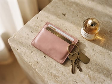 Flatlay of a soft pink leather cardholder, keys, and a small perfume bottle on a beige stone surface, soft shadows, warm morning light, 35mm