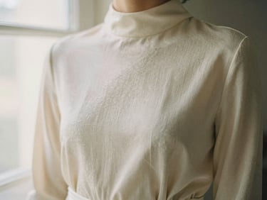A high-neck long-sleeve dress in textured cream silk, softly lit by diffused window light, focusing on the fabric texture.