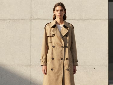 A tall model in a structured tan trench coat standing against a minimalist concrete wall, soft warm afternoon sun.