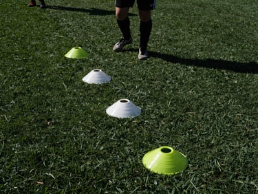 Wide panoramic shot, low angle on a player's feet executing a rapid cone drill on turf, high-contrast natural midday sun, harsh shadows.