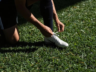 Candid sideline moment of a player lacing up cleats, deep shadows, natural light.