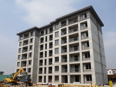 Wide shot of a completed residential apartment complex exterior, natural daylight, multiple floors with balconies and structural concrete framing visible, construction equipment parked at the base for scale, overcast sky