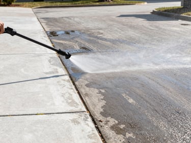 Pressure washer spraying a concrete driveway in Columbus Georgia, a clear line between clean white concrete and grimy gray surface, water mist catching the midday sunlight, wide framing showing the full driveway width
