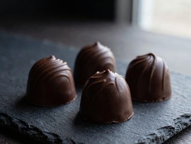 Macro shot of three glossy, hand-painted artisanal chocolate bonbons on a dark slate surface, dramatic side lighting.