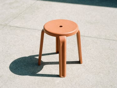 Low-angle architectural shot of a single terracotta-colored resin stool casting a sharp shadow on raw concrete under harsh midday sun, medium format film