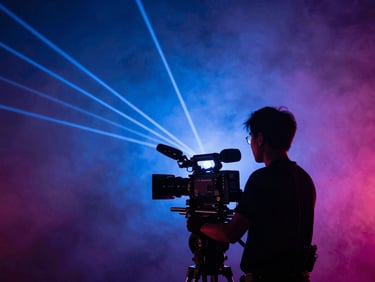 Tight telephoto shot of a videographer in silhouette operating a cinema camera on a shoulder rig, dramatic stage lighting behind, laser beams cutting through smoke, rich blue and magenta tones, shallow focus on the camera lens, documentary style