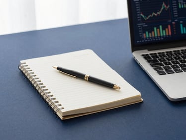 Clean minimalist workspace with a notebook, a pen, and a laptop displaying financial metrics, daylight