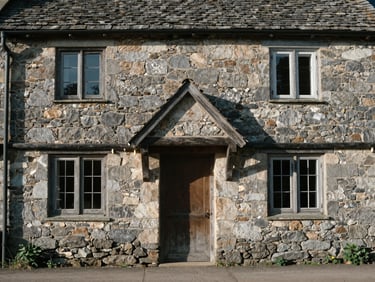 Straight-on architectural photograph of a stone cottage, original timber lintels, soft daylight, low-contrast shadows.