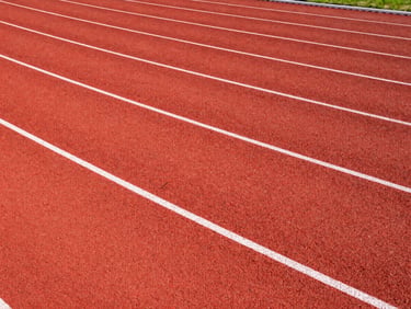 High-contrast architectural shot of a red polyurethane athletic track, crisp white lane markings, wide-angle lens, bright crisp daylight.
