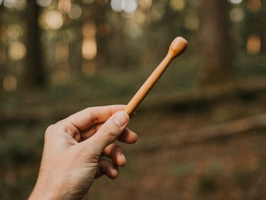 A hand holding a simple wooden kuripe applicator, detailed texture of the wood, soft natural forest background, warm lighting
