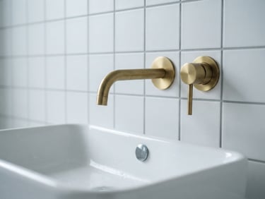 Close-up of a modern wall-mounted brass tap over a white ceramic basin, clean grey grout lines, bright daylight