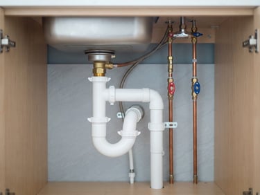 Neat under-sink plumbing with white waste pipes and copper hot-cold lines under a kitchen counter, clean layout