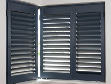Modern bedroom window with motorized dark gray aluminum shutters partially open, soft natural light filtering through, sharp clean lines.