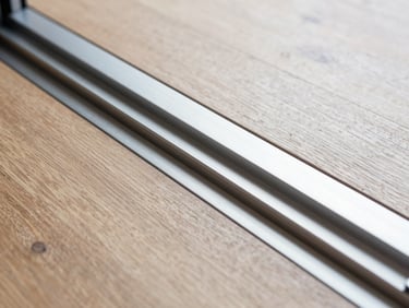Macro architectural shot of a recessed minimalist aluminum sliding door track flush with hardwood flooring, clean geometric lines, metallic texture, bright daylight.