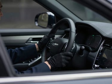 Interior shot of a luxurious, dark-tinted SUV, focusing on the steering wheel and dashboard, with a glimpse of an operative's gloved hand on the wheel, cinematic editorial photography, low-key lighting, muted navy and charcoal tones, sharp focus, professional and discreet atmosphere, 35mm