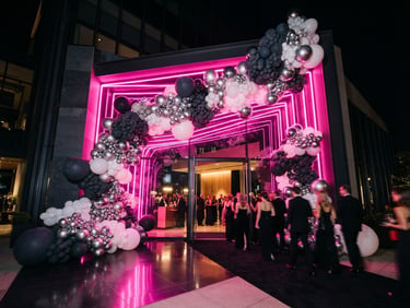 Luxury event entrance with an asymmetrical architectural balloon installation in charcoal, white, and silver, backlit by intense pink neon light strips, professional editorial photography, wide-angle 35mm.