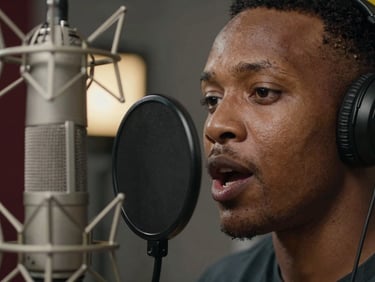 Close-up of a voice actor mid-take at a microphone in a dim studio, mouth slightly open mid-word, north-facing daylight on one cheek, warm lamp glow behind, shallow depth of field, Addis Ababa studio environment