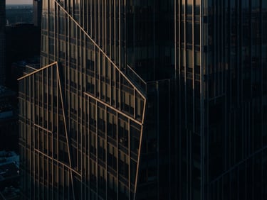 Moody corporate photography of abstract architectural lines of a modern building facade, soft directional window light with deep shadows, deep navy and warm gold tones, 35mm